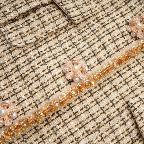 Woman wearing gold tweed sequin jacket and matching skirt set by Aelle standing against plain background