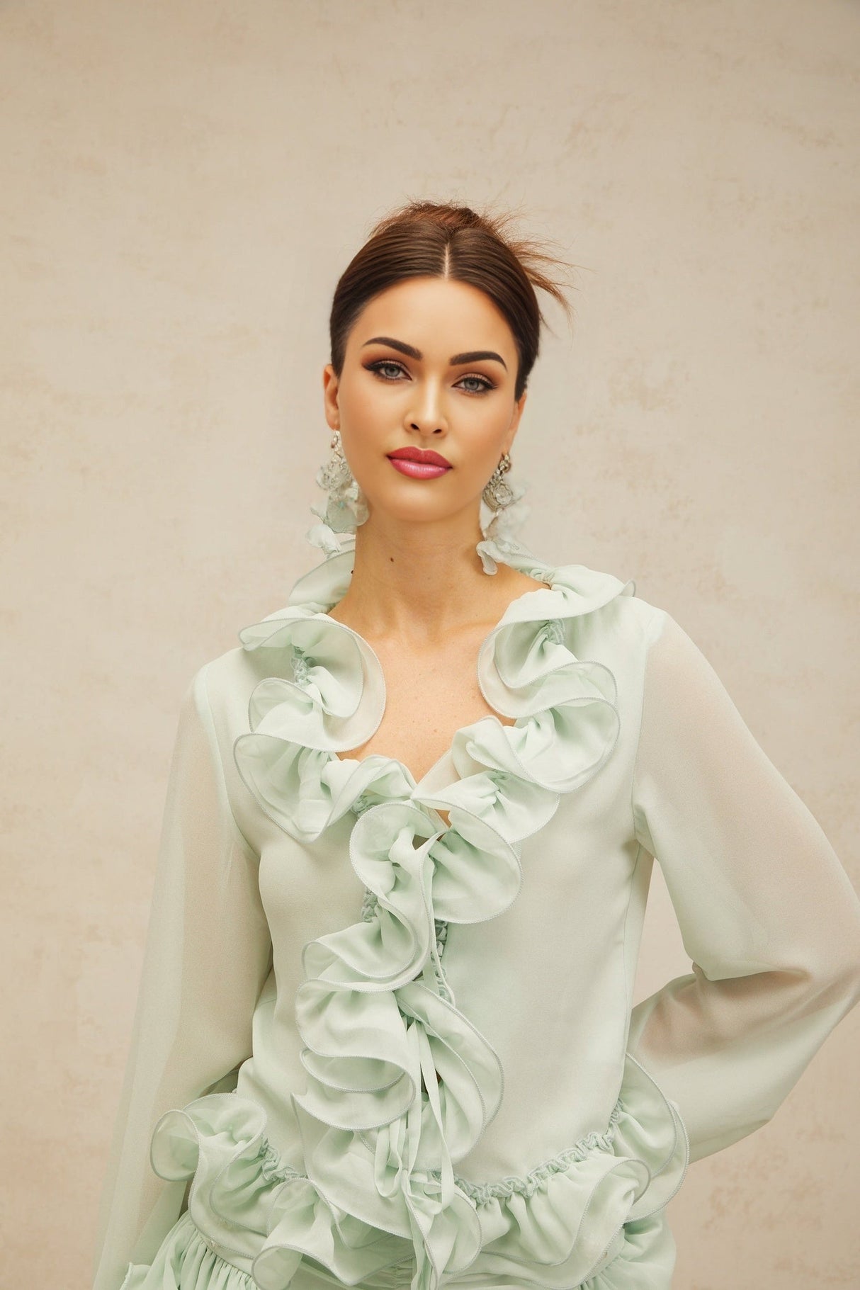 Woman wearing green chiffon blouse with ruffled details and matching flowy skirt standing against neutral background