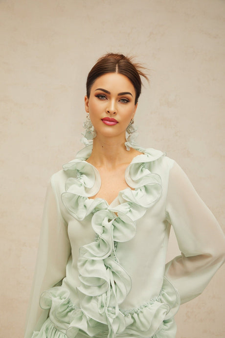 Woman wearing green chiffon blouse with ruffled details and matching flowy skirt standing against neutral background