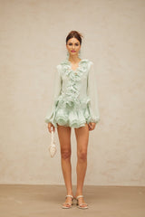 Woman wearing green chiffon blouse with ruffled details and matching flowy skirt standing against plain background
