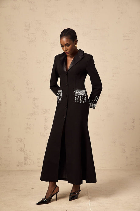 Woman wearing long black blazer dress with embellishments standing against plain background