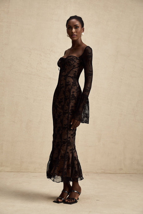 Woman wearing long black flared lace maxi dress with Marlene style silhouette standing against plain background