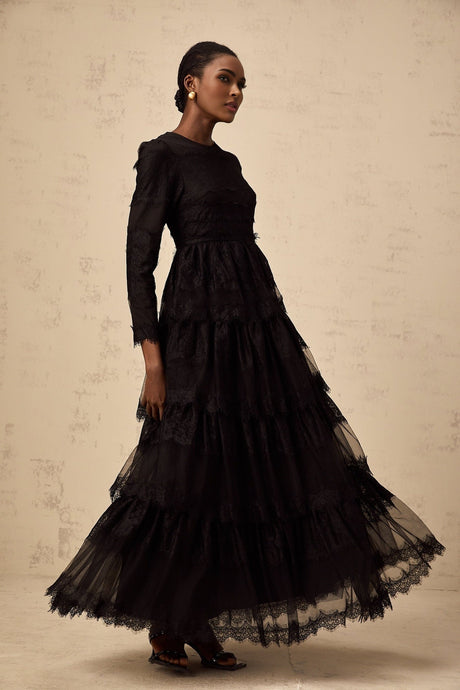 Woman wearing long black lace dress with tiered skirt and sheer tulle accents standing against plain background