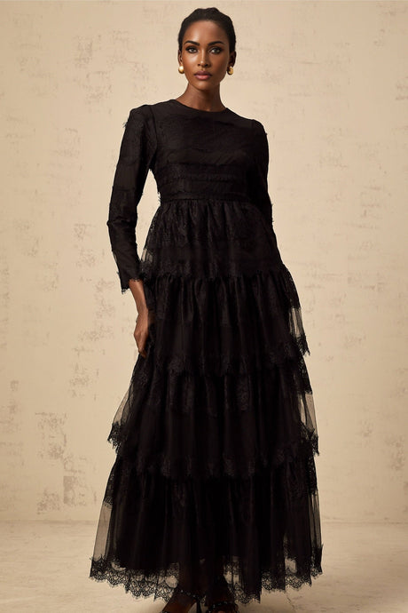 Woman wearing long black lace dress with tiered skirt and sheer tulle details standing against plain background