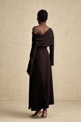Woman wearing long black pleated maxi dress with high slit posing against plain background