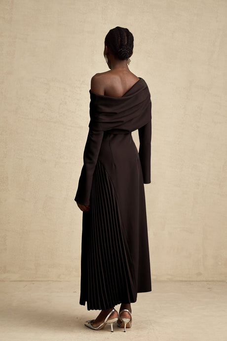 Woman wearing long black pleated maxi dress with high slit posing against plain background