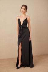 Woman wearing long black satin maxi dress with V neckline and front slit standing against plain background