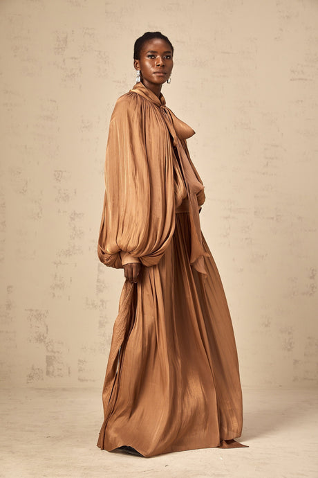 Woman wearing long brown pleated satin maxi dress with flowing skirt and fitted bodice
