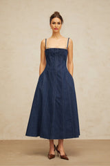 Woman wearing long denim dress with panel detailing in dark blue shade standing against plain background