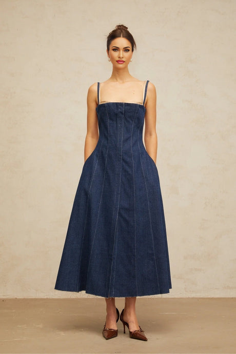 Woman wearing long denim dress with panel detailing in dark blue shade standing against plain background