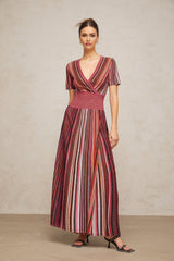 Woman wearing long flowing metallic knit maxi dress with multicolor stripes in shimmering tones standing against plain background