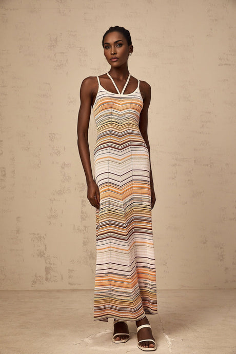 Woman wearing long striped knit maxi dress with multicolored horizontal stripes in pastel shades standing against neutral background