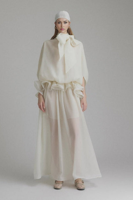 Woman wearing long white flowing maxi dress with ruffled details and tie neck standing in sunlit outdoor setting