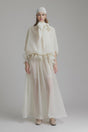 Woman wearing long white flowing maxi dress with ruffled details and tie neck standing in sunlit outdoor setting