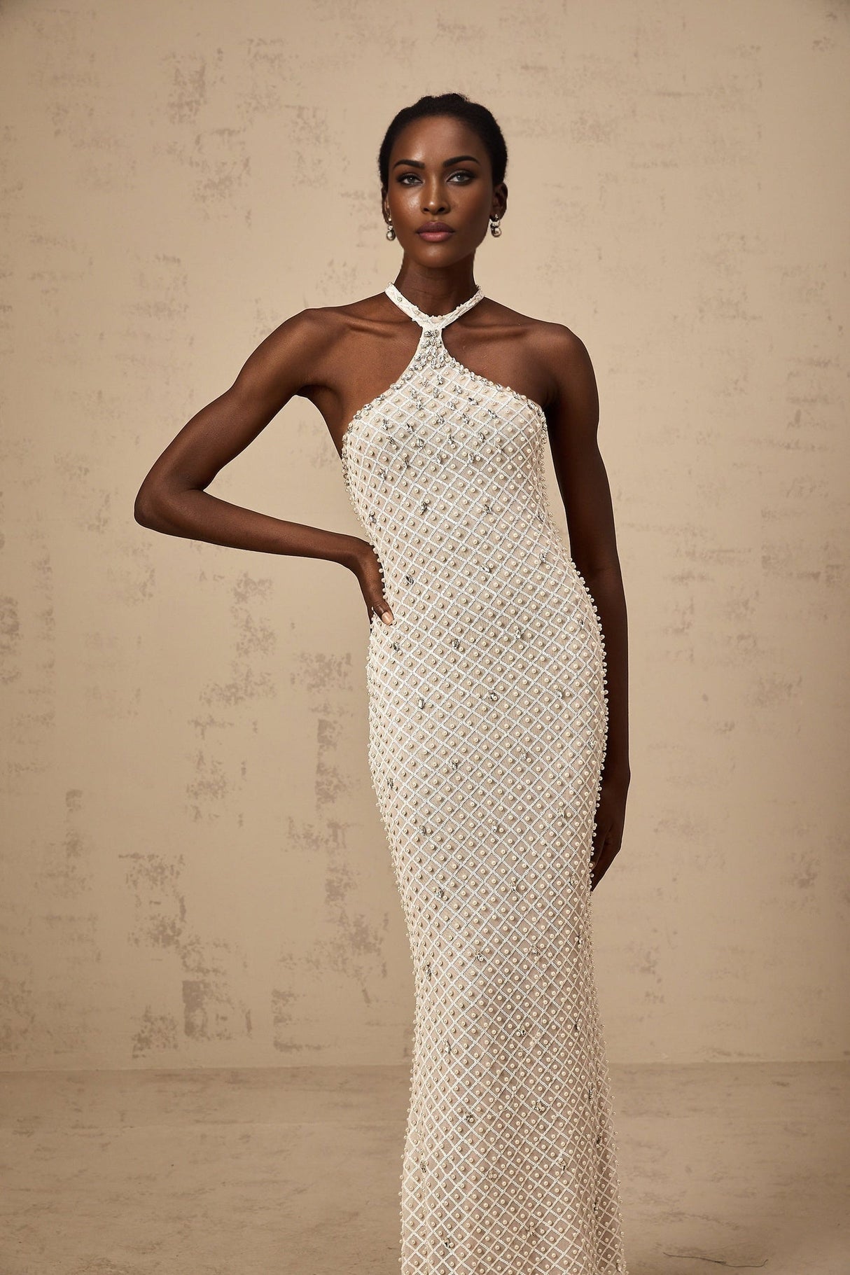 Woman wearing long white halterneck maxi dress with embellished details standing against plain background