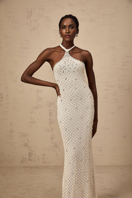 Woman wearing long white halterneck maxi dress with embellished details standing against plain background