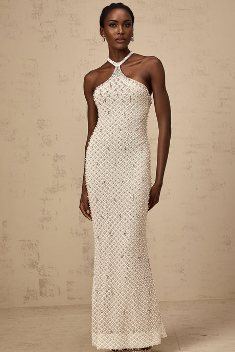 Woman wearing long white halterneck maxi dress with embellished details standing against plain background