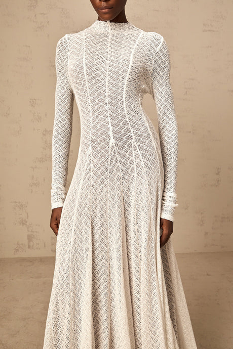 Woman wearing long white lace maxi dress with wavy pattern and sleeveless design standing against plain background