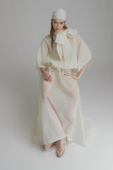 Woman wearing long white maxi dress with ruffled details and tie neck standing in outdoor setting