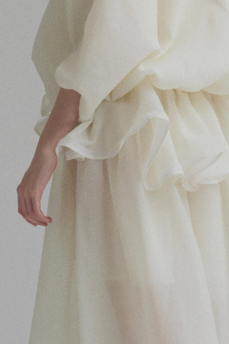 Woman wearing long white maxi dress with tie neck and ruffled details standing against plain background