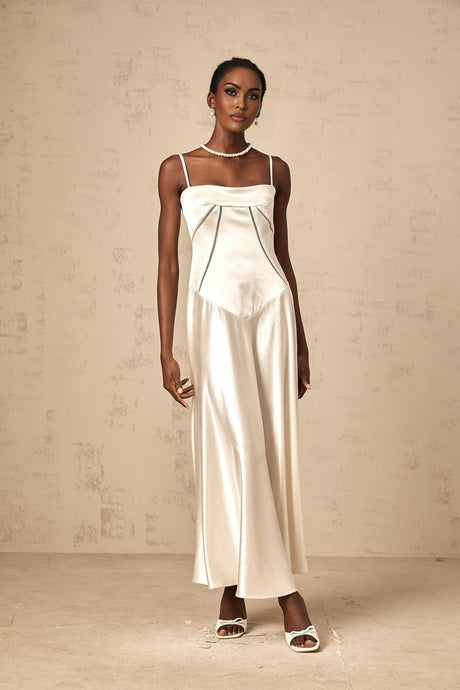 Woman wearing long white satin maxi dress with flared skirt and sleeves standing against plain background