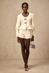 Woman wearing matching crystalembellished jacket and shorts set in light color fabric with sparkly details standing against plain background