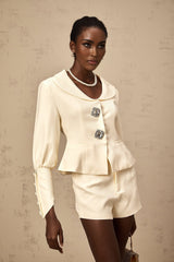 Woman wearing matching crystalembellished jacket and shorts set in light color standing against plain background