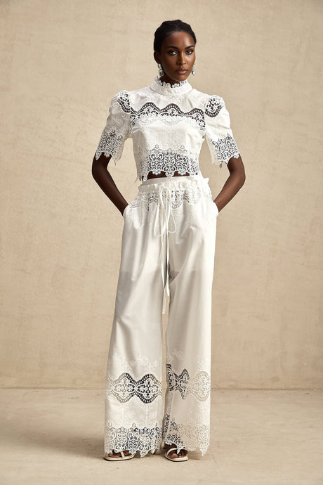 Woman wearing matching white lace trimmed cropped top and trousers set with cutout details
