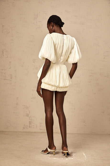 Woman wearing short beige dress with pleated skirt and floral appliques standing against neutral background