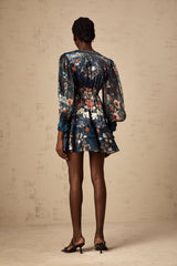 Woman wearing short blue dress with graphic print pattern and embellishments standing in relaxed pose against plain background