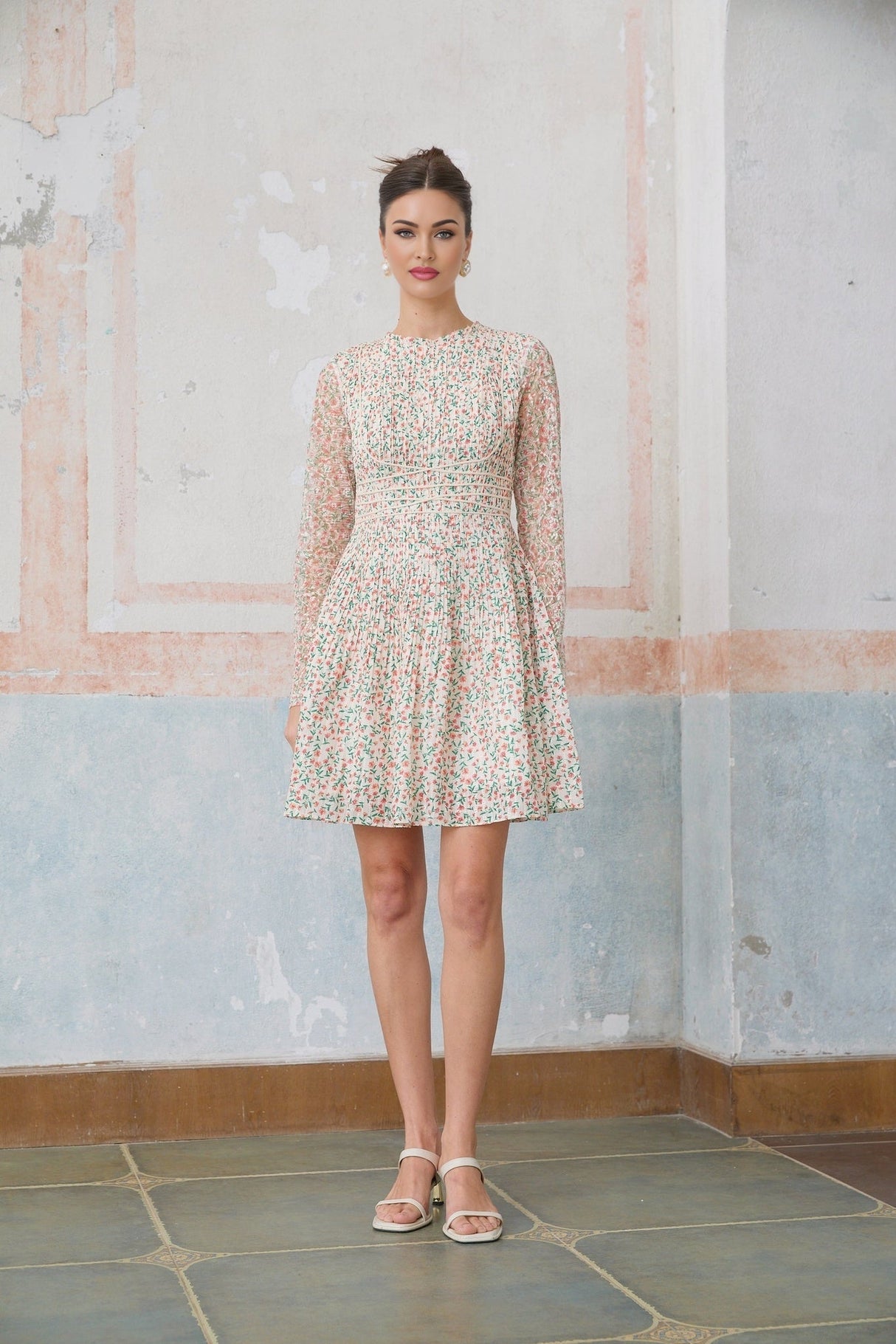 Woman wearing short floral print dress with pleated skirt and Marion design standing against plain background
