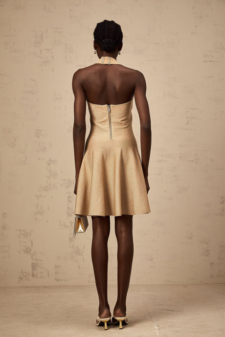 Woman wearing short khaki knitted dress with embellishments and long sleeves standing against plain background