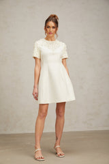 Woman wearing short white dress with colorful floral appliques standing against plain background