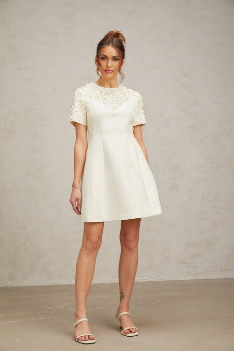 Woman wearing short white dress with colorful floral appliques standing against plain background