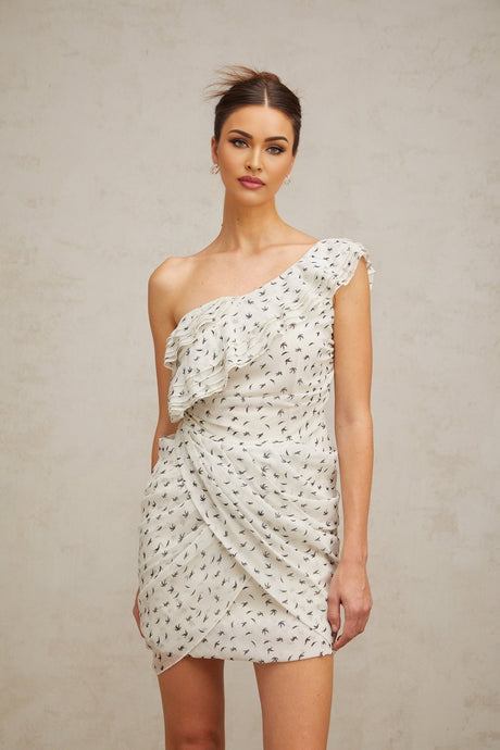 Woman wearing short white dress with ruffles and printed pattern standing against plain background