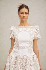 Woman wearing short white lace dress with puffed sleeves and floral pattern standing against plain background