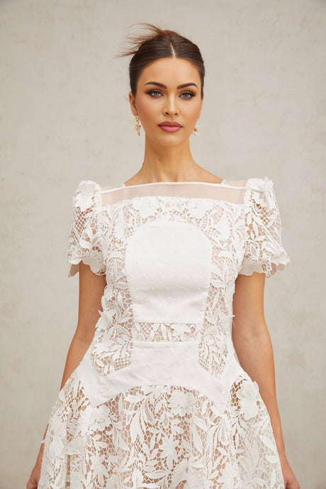 Woman wearing short white lace dress with puffed sleeves and floral pattern standing against plain background