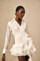 Woman wearing short white semi sheer dress with floral pattern and ruffled details standing against plain background