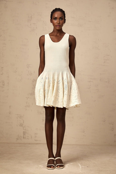 Woman wearing short white textured knit dress with intricate pattern standing against plain background