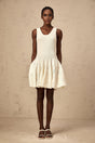 Woman wearing short white textured knit dress with intricate pattern standing against plain background