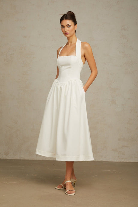 Woman wearing white halterneck midi dress with pleated skirt and fitted bodice standing against plain background