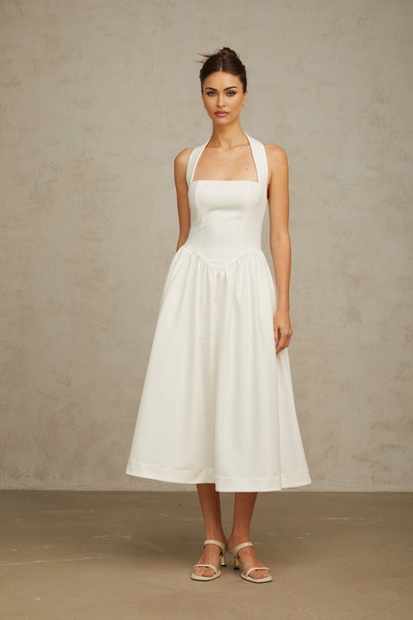 Woman wearing white halterneck pleated midi dress with flowy skirt standing against plain background