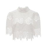 Woman wearing white lace trim cropped top and matching trousers with cutout details standing against plain background