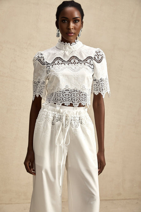 Woman wearing white lace trimmed cropped top and matching trousers with cutout details Raphaelle set