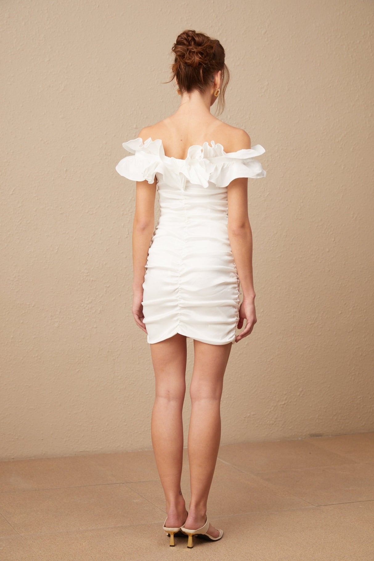 Woman wearing white off shoulder mini dress with ruffled details standing against plain background