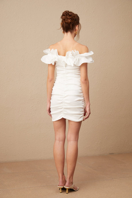 Woman wearing white off shoulder mini dress with ruffled details standing against plain background