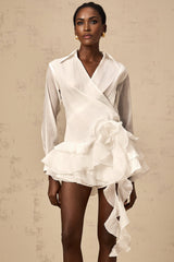 Woman wearing white semi sheer mini dress with floral pattern and ruffled details standing against plain background