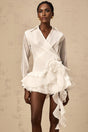 Woman wearing white semi sheer mini dress with floral pattern and ruffled details standing against plain background
