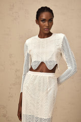 Woman wearing white twopiece outfit consisting of a wavy woven top and matching skirt standing against plain background