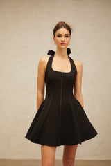 Young woman wearing a black halterneck mini dress with bow detail at the neck standing against a plain background